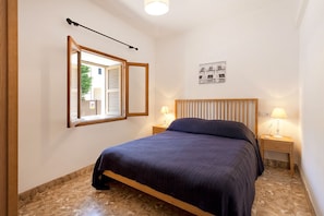 Een slaapkamer met een bed, nachtkastje met lamp, raam met jaloezieën en een spiegel aan de muur.