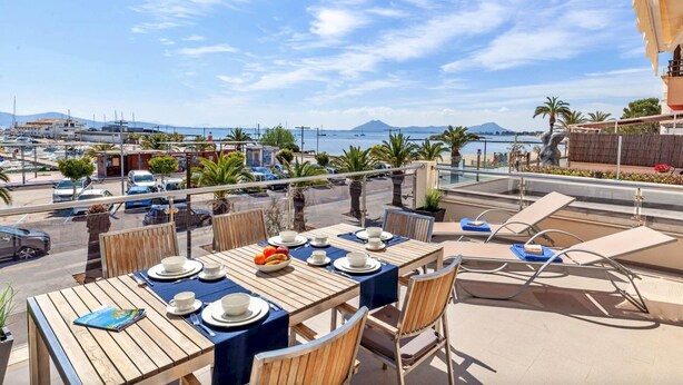 Un espace de restauration en plein air avec des tables pour deux, offrant une vue sur une marina avec des bateaux et une montagne au loin.