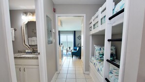Un pasillo con un baño a la izquierda, que incluye un lavabo y un espejo. A la derecha, hay literas con ropa de cama estampada. Una puerta conduce a una sala de estar con vistas al exterior.