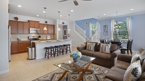 A modern kitchen with a central island, a dining area with a staircase, and a living room with a sofa and coffee table. - The Majorca at Windsor Westside (Kissimmee)