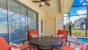 A covered patio with a pool table, four orange chairs with floral cushions, and a ceiling fan. - The Majorca at Windsor Westside (Kissimmee)