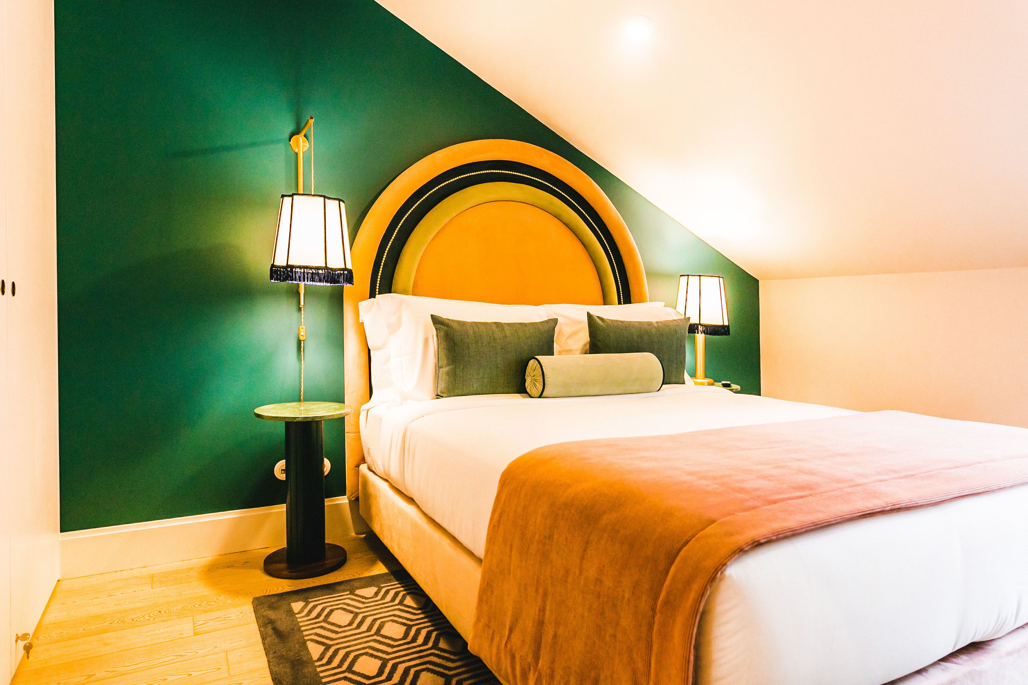 Habitación de hotel con una cama, dos lámparas de noche, una pared de acento verde y una alfombra estampada.