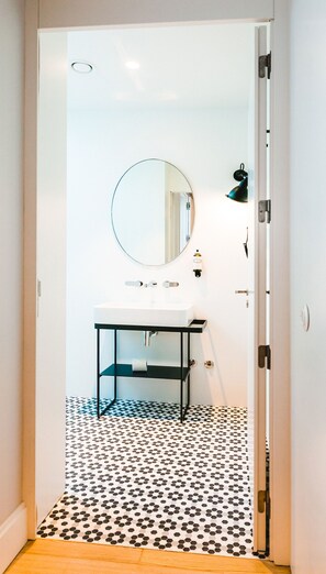 A bathroom with a white sink, a round mirror, and a black metal shelf. The floor has a black and white polka dot tile pattern. - The Lift Apartments by RIDAN Hotels (Lisbon)