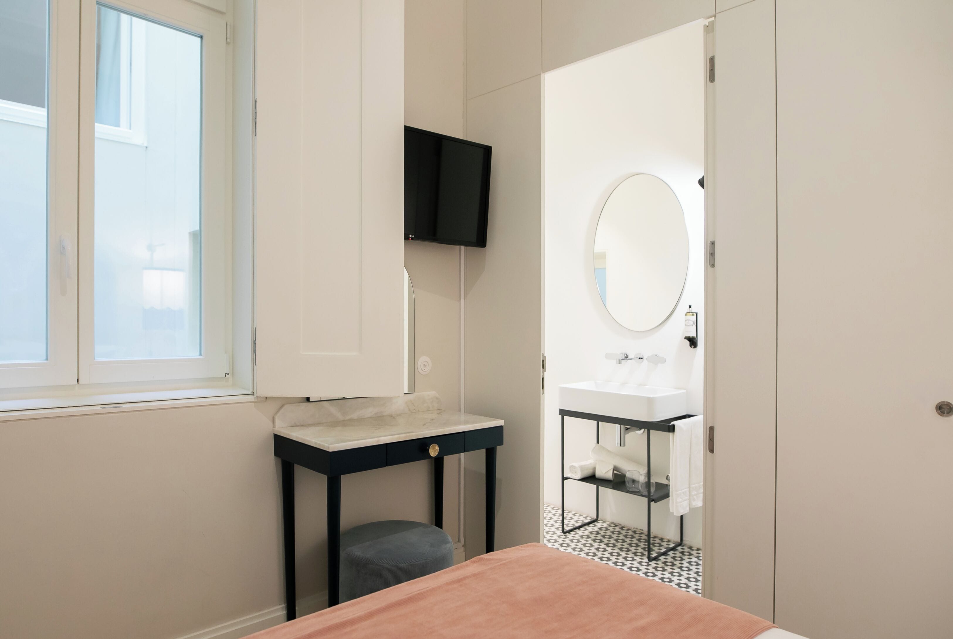 Un dormitorio con cama, mesita de noche, televisor montado en la pared y baño visible a través de una puerta abierta.