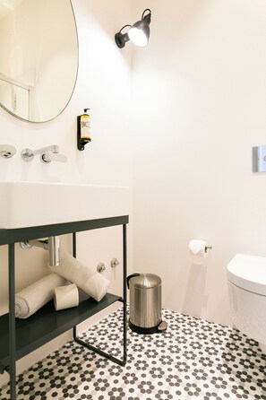 A bathroom with a white sink, a mirror, a towel rack, a toilet paper holder, a trash can, and a floor with a black and white hexagonal pattern. - The Lift Apartments by RIDAN Hotels (Lisbon)