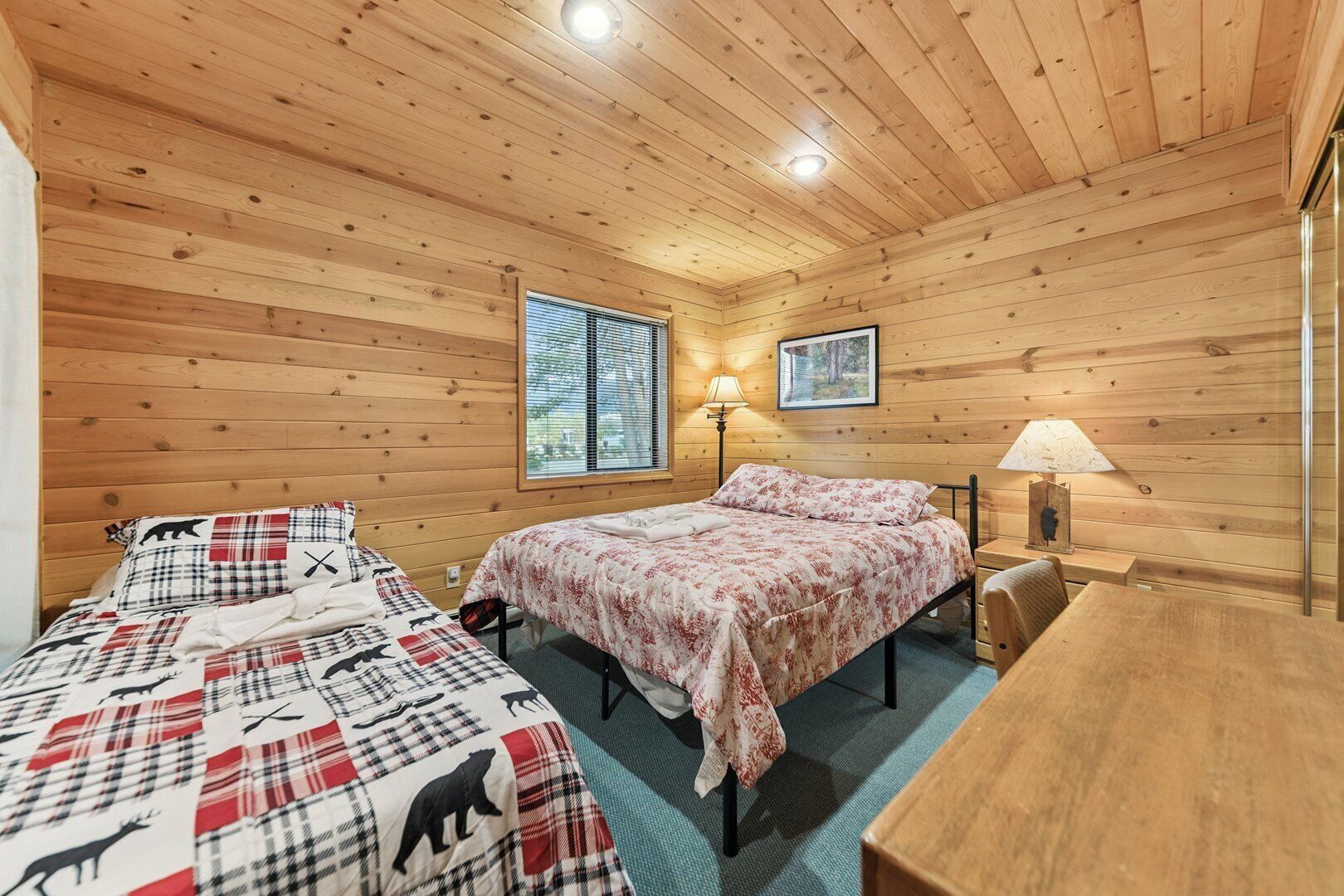 Una habitación acogedora tipo cabaña con dos camas, techo de madera, ventana con persianas, un cuadro en la pared y una mesa con lámpara.