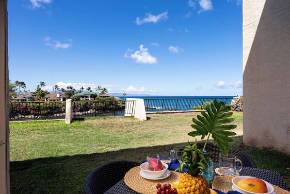 A table with tropical fruits and a drink, overlooking a beach with palm trees and houses.