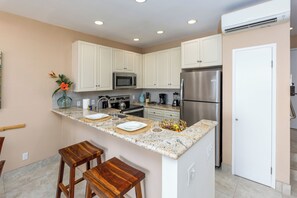 A modern kitchen with white cabinets, a stainless steel refrigerator, a microwave, and a coffee maker. There are two wooden stools and a vase with flowers on the island.