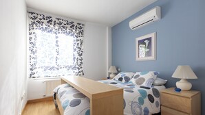 Un dormitorio con cama, mesitas de noche, ventana con cortinas florales, unidad de aire acondicionado y un cuadro en la pared.