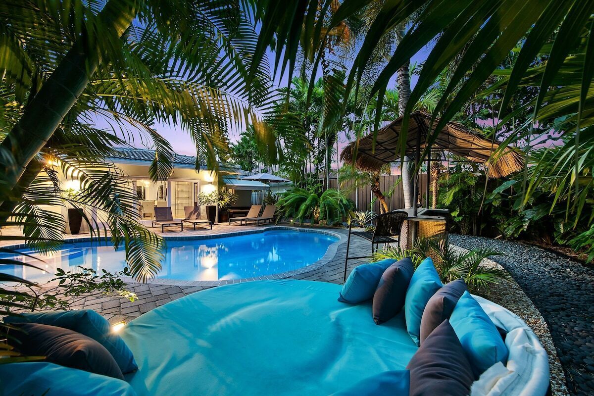 A swimming pool with lounge chairs and pillows, surrounded by tropical plants and a thatched-roof structure.