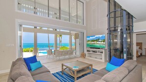 A spacious living room with a large sofa, a coffee table, and a television. There is a glass elevator and a view of the ocean through large windows. - Abita Kai: ADA-Friendly Villa Walkable to Restaurants, w/Pool, Dock, & Sandy Beach (Grand Cayman)