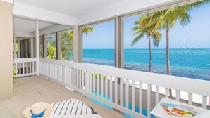A balcony with a view of the ocean and palm trees, a white railing, and a table with a menu. - Abita Kai: ADA-Friendly Villa Walkable to Restaurants, w/Pool, Dock, & Sandy Beach (Grand Cayman)
