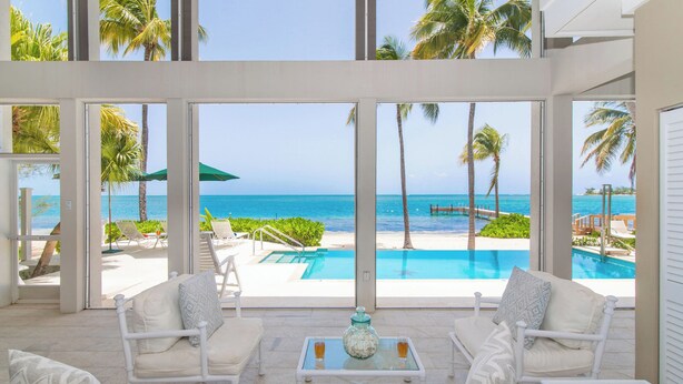 A room with a view of a pool and beach, white furniture, and a glass table with a jar and glasses. - Abita Kai: ADA-Friendly Villa Walkable to Restaurants, w/Pool, Dock, & Sandy Beach (Grand Cayman)