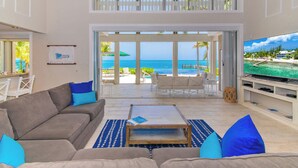 A spacious living room with a large flat-screen TV, a comfortable sofa, and a view of the ocean through large glass doors. - Abita Kai: ADA-Friendly Villa Walkable to Restaurants, w/Pool, Dock, & Sandy Beach (Grand Cayman)