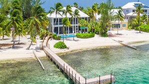 A coastal property with a wooden dock extending into clear turquoise water, surrounded by palm trees and a large building with a pool. - Abita Kai: ADA-Friendly Villa Walkable to Restaurants, w/Pool, Dock, & Sandy Beach (Grand Cayman)