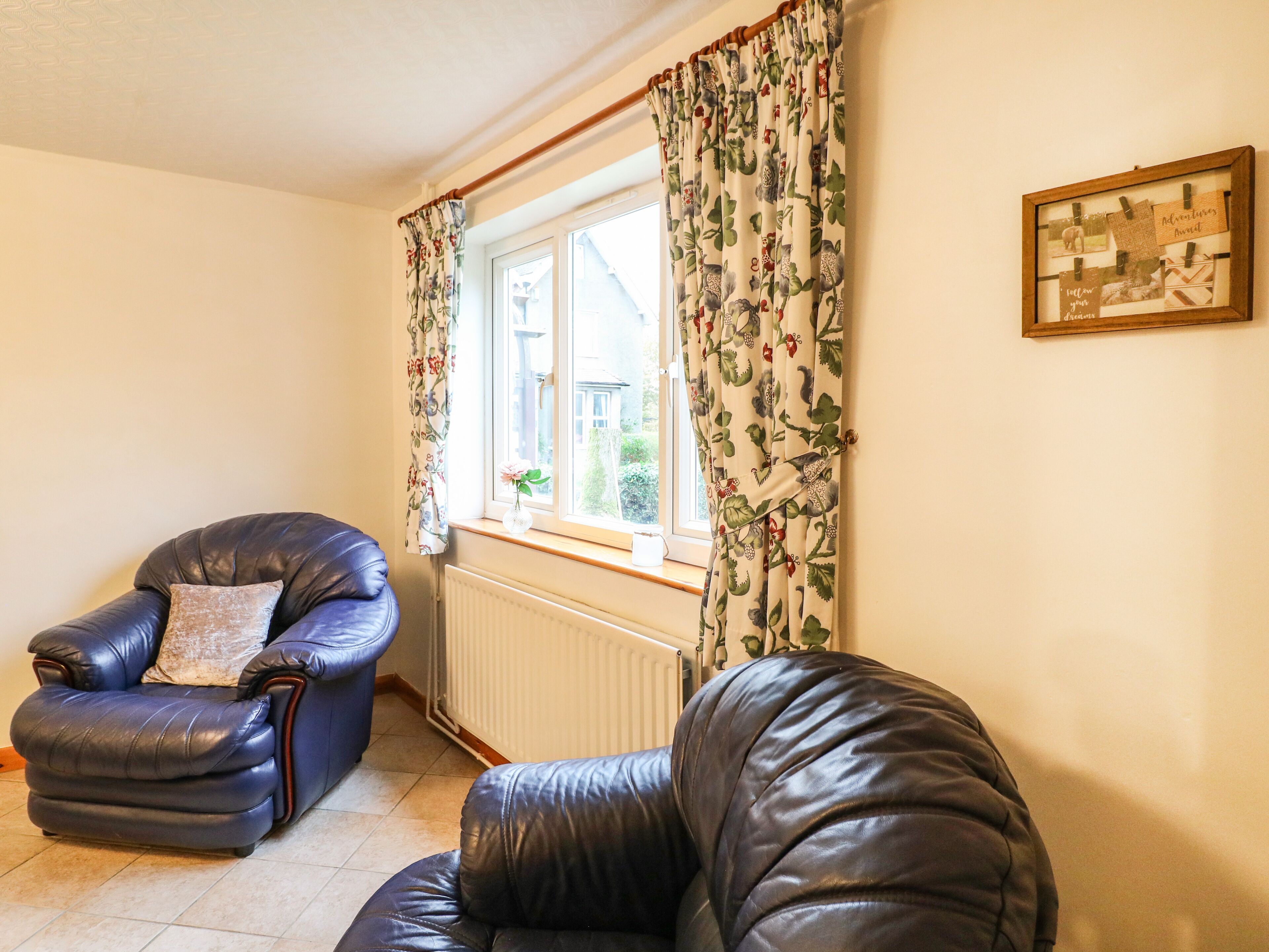 A living room with two leather armchairs, a radiator, a window with floral curtains, and a framed picture on the wall.