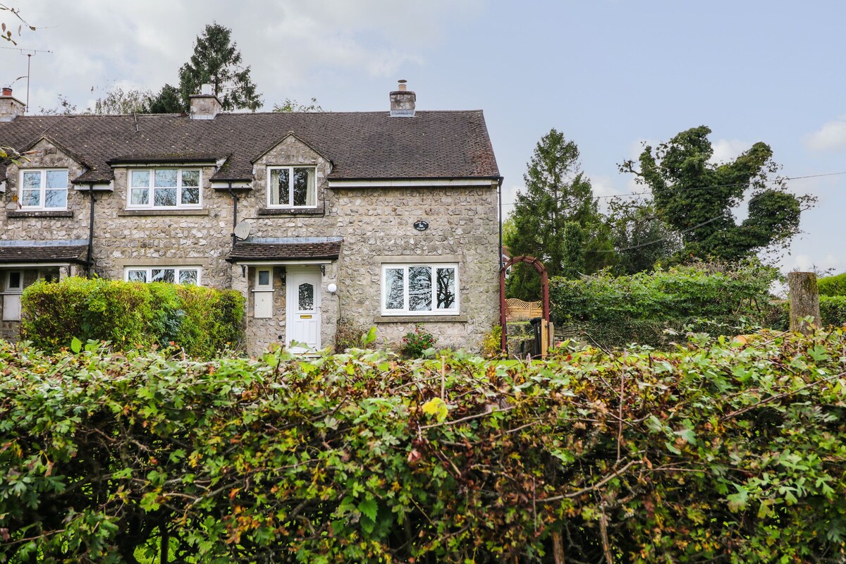 A stone house with a white door and multiple windows, surrounded by dense greenery.