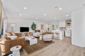 A modern living room with a sofa, coffee table, and TV. There is a kitchen area with white cabinets and a dining table.