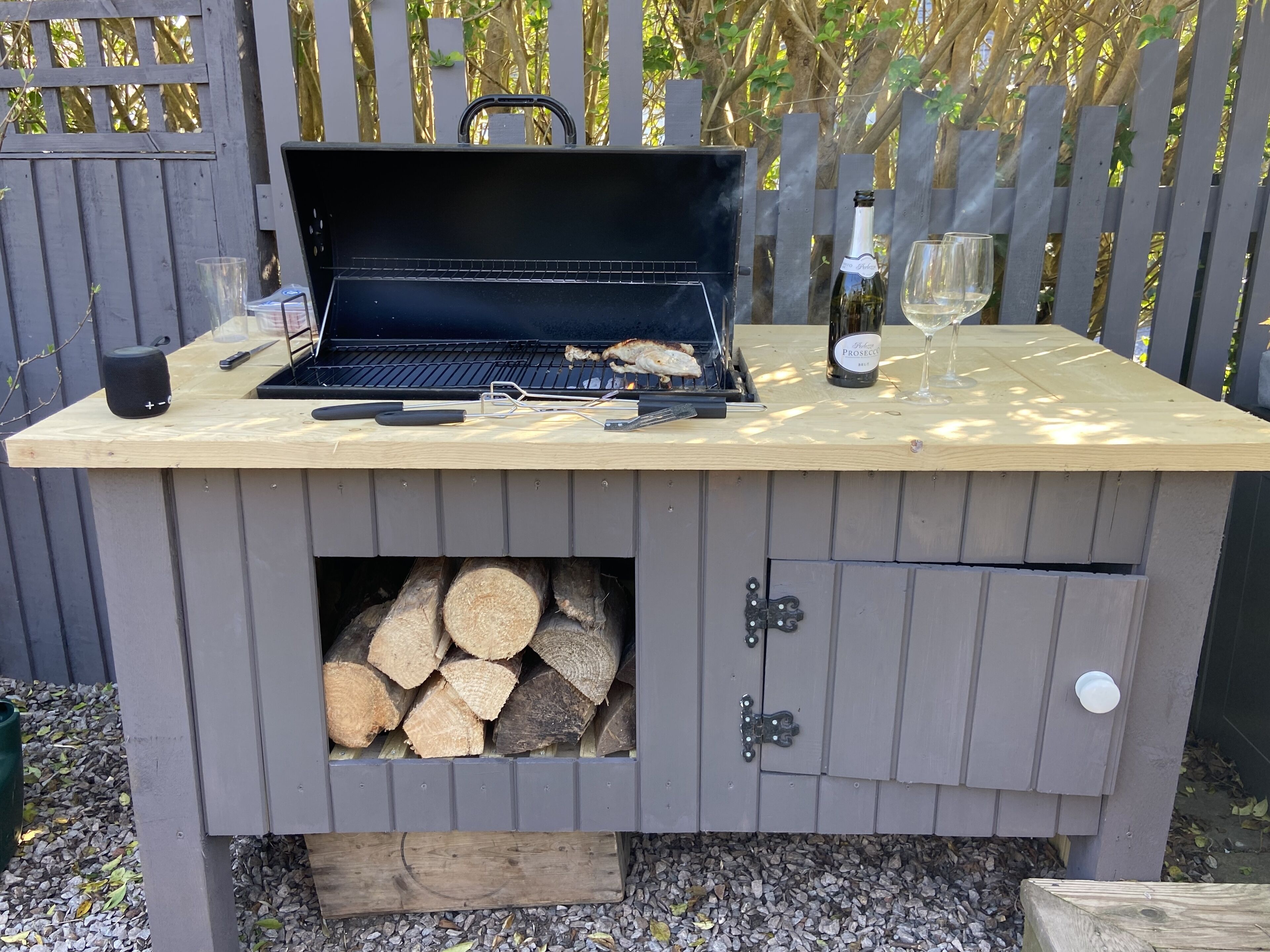 Una griglia per barbecue in legno con un vano portaoggetti integrato per legna, una bottiglia di vino e un bicchiere sulla superficie della griglia.