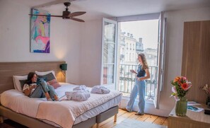 A bedroom with a bed, a ceiling fan, a window with a view of buildings, and a vase with flowers. - LOLA Boutique Hôtel Bordeaux Centre (Bordeaux)