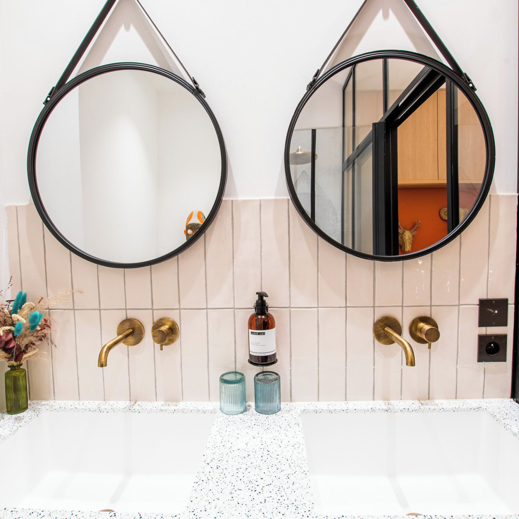 Un baño con dos espejos redondos, una pared de azulejos blancos, un lavabo con detalles de latón y un jarrón pequeño con flores secas.