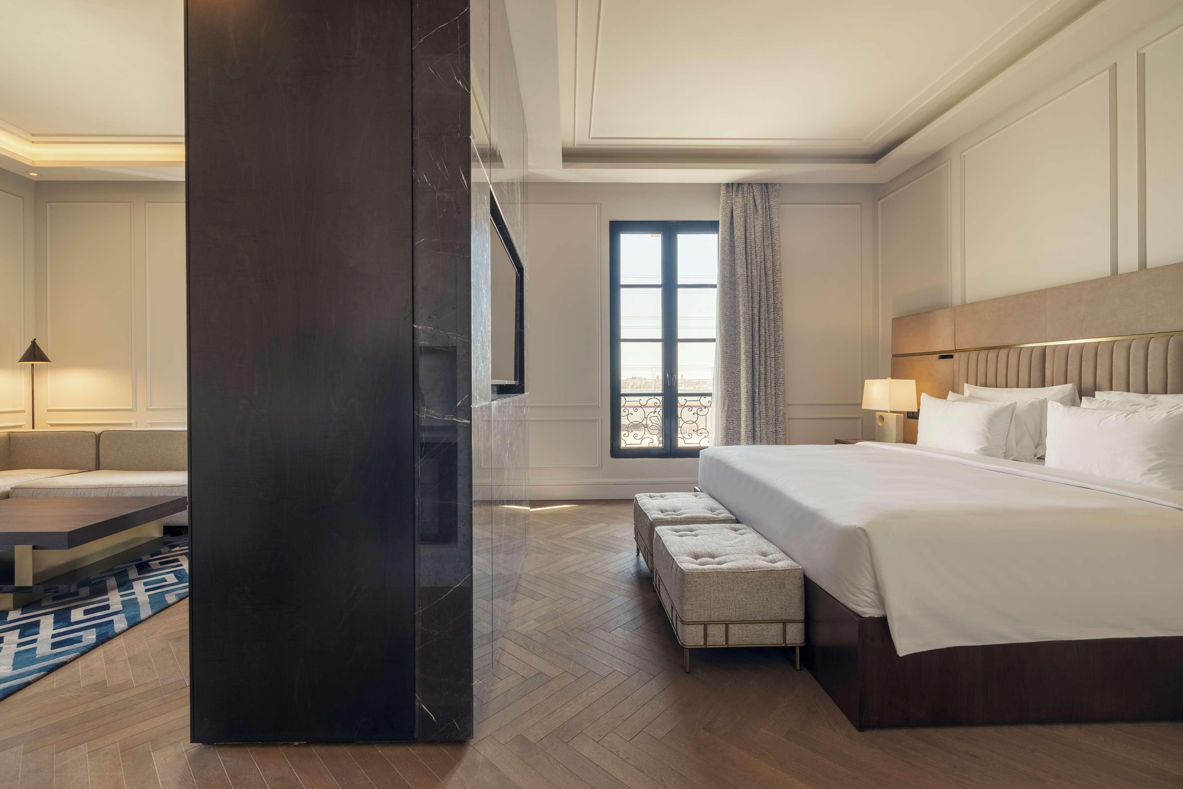 Ein Hotelzimmer mit einem großen Bett, einem Schreibtisch, einem Stuhl, einem Fenster mit Vorhängen und Blick auf die Stadt.