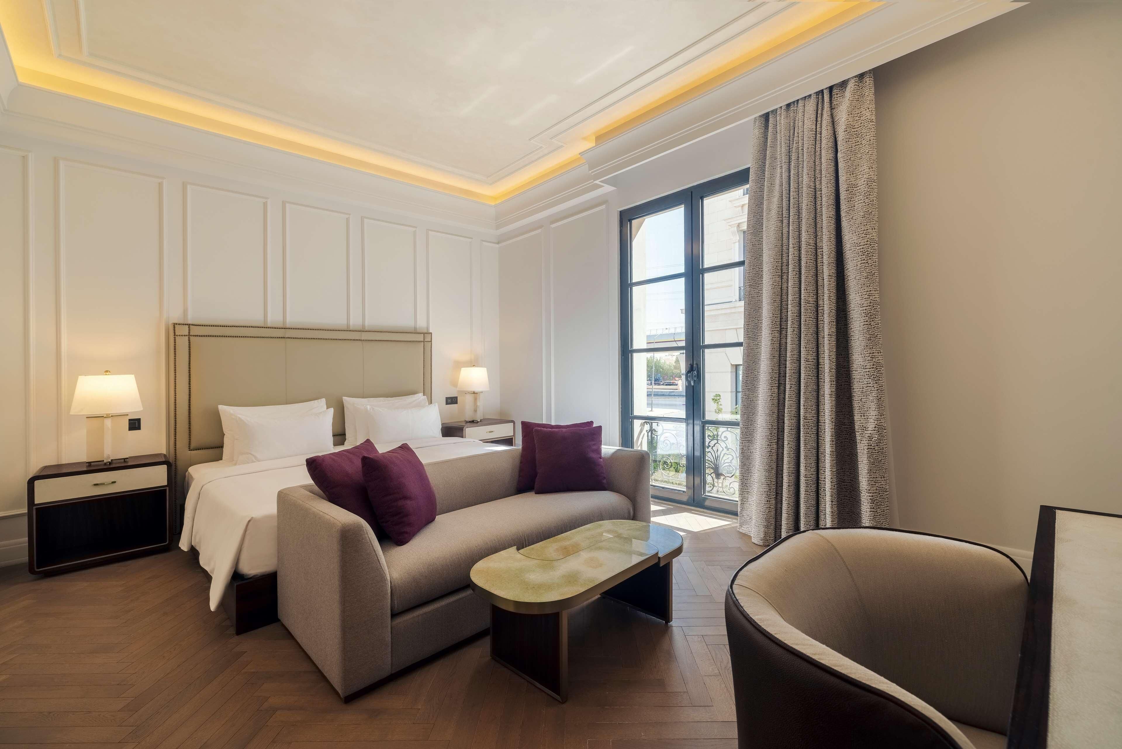 Ein Hotelzimmer mit Bett, Sofa, Sessel, Nachttisch und einem Fenster mit Vorhängen.