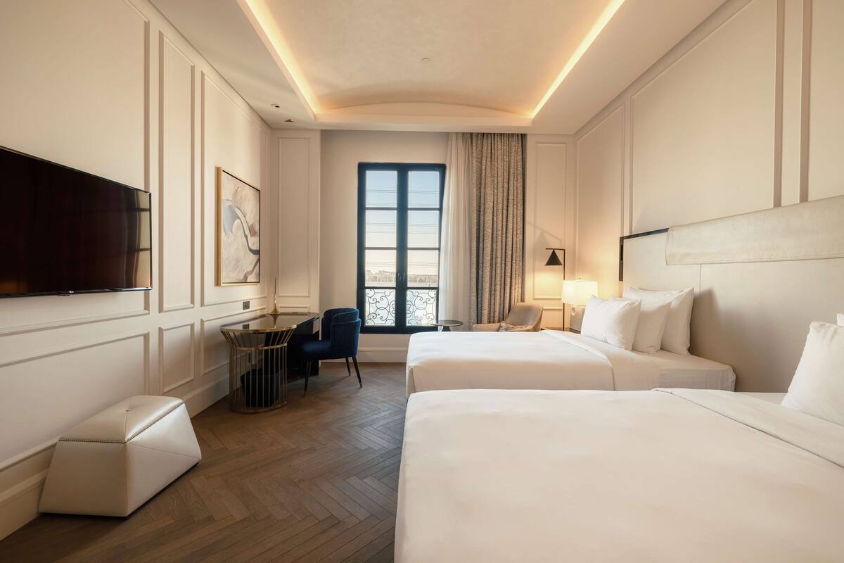 A hotel room with two beds, a desk, a chair, a TV, and a window with curtains.