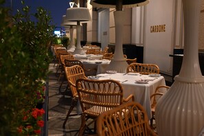 An outdoor dining area with wicker chairs and white tablecloths, featuring a 'CARBONE' sign on the wall.