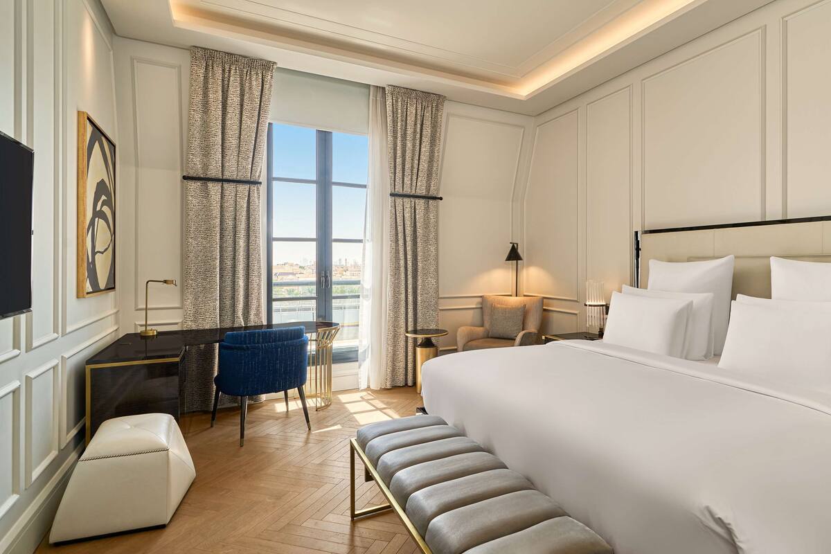 A hotel room with a large bed, a desk, a chair, a small table, and a TV.