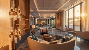 Un hall amplio con diseño moderno, que cuenta con una zona de estar central con sofás y sillones, una lámpara de araña y ventanas grandes con vistas al exterior.