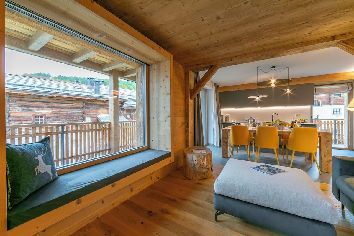 A wooden cabin interior with a dining area, wooden bench with a cushion, and a view through a window.