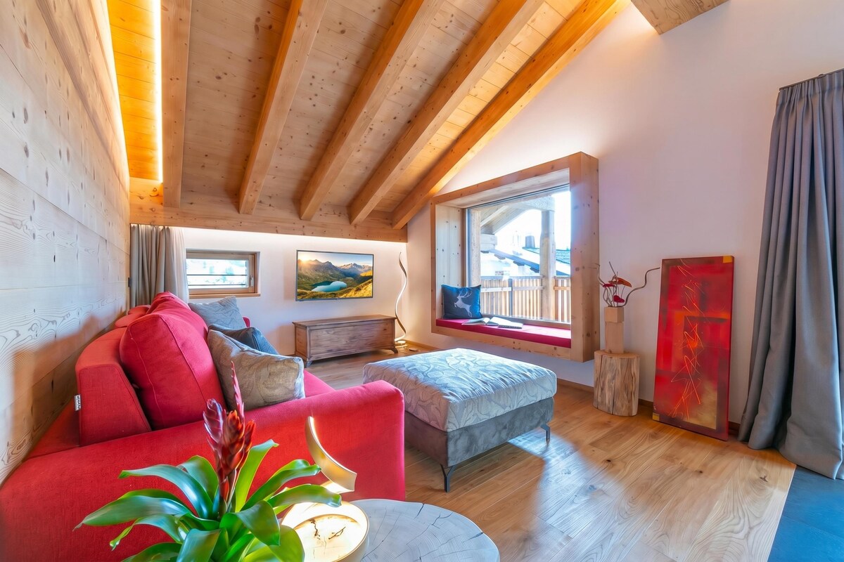A cozy room with a red sofa, a small round table with a plant, and a window with a view of mountains.