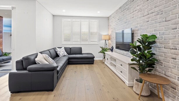 A modern living room with a sectional sofa, a flat-screen TV mounted on a brick wall, and a wooden floor. - Carrington Nelson Bay (Nelson Bay)