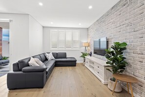 Ein modernes Wohnzimmer mit einer Eckcouch, einem Flachbildfernseher an einer Ziegelwand und Holzdielenboden.