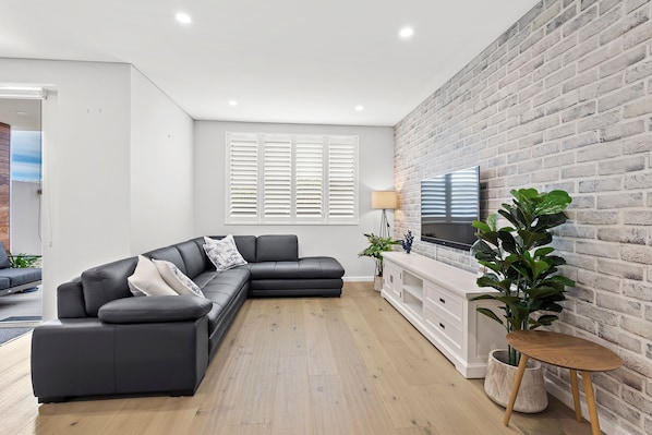 Un salon moderne avec un canapé d’angle, un téléviseur à écran plat fixé sur un mur en briques et un plancher en bois.