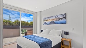 A bedroom with a bed, a nightstand, a lamp, a painting, and a sliding glass door leading to an outdoor area with plants. - Carrington Nelson Bay (Nelson Bay)