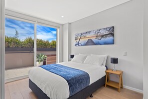 Une chambre à coucher comprenant un lit, une table de chevet, une lampe, un tableau et une porte-fenêtre coulissante donnant sur un espace extérieur agrémenté de plantes.