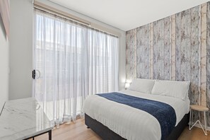 Ein Schlafzimmer mit einem Bett, einem Nachttisch, einer Lampe und Blick ins Freie durch transparente Vorhänge.