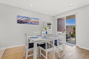 Un coin repas avec une table et des chaises blanches, un tableau accroché au mur et une vue sur l’extérieur, visible à travers une porte ouverte.