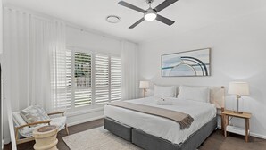 A bedroom with a bed, a ceiling fan, a window with blinds, a bedside table with a lamp, and a chair. - Maggie Mae Family Retreat in Shoal Bay with Pool (Shoal Bay)