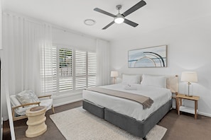 A bedroom with a bed, a ceiling fan, a window with blinds, a bedside table with a lamp, and a chair. - Maggie Mae Family Retreat in Shoal Bay with Pool (Shoal Bay)