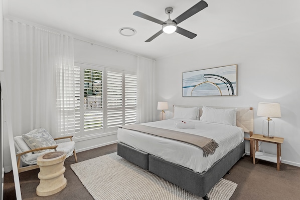 A bedroom with a bed, a ceiling fan, a window with blinds, a bedside table with a lamp, and a chair.