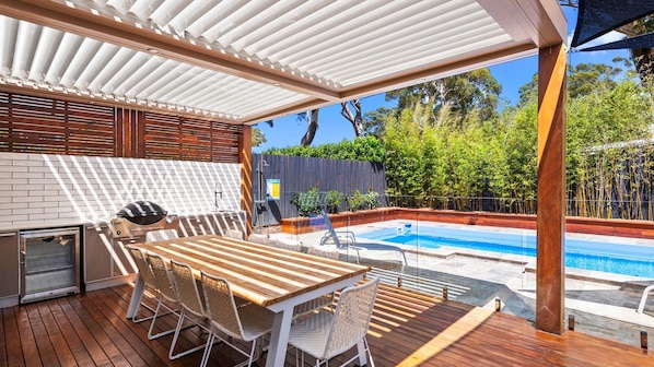 An outdoor patio with a wooden table and chairs, a barbecue grill, a pool, and a covered area with a white roof. - Ultimate family holiday in Style (Shoal Bay)