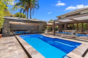 A swimming pool with clear blue water, surrounded by stone tiles. There are lounge chairs and a glass-enclosed area with a thatched roof.