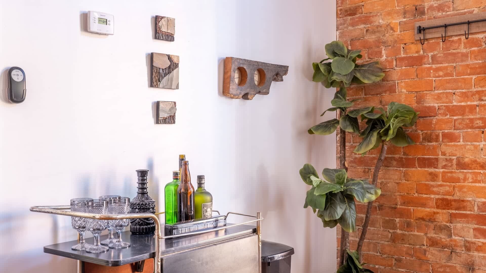 Un espace bar avec un chariot en métal servant de présentoir pour les verres et les bouteilles, un mur en briques et une plante suspendue.