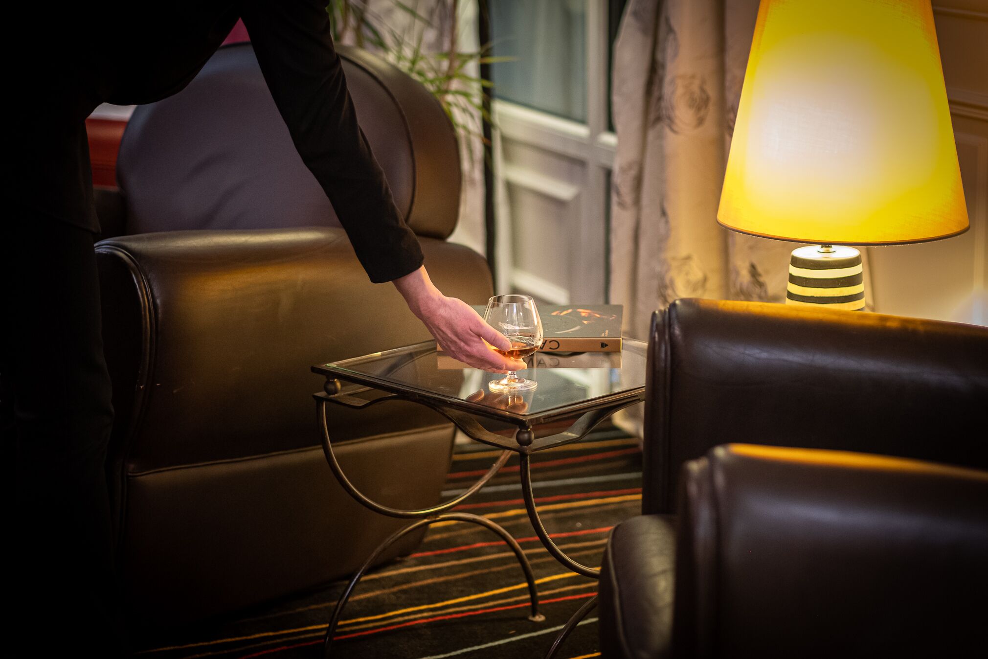 Eine Person stellt ein Glas auf einen Glastisch neben eine Couch in einem Zimmer mit einer Lampe.
