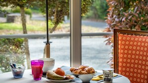 Ein Frühstückstisch mit Croissants, einer Schale Joghurt und einem Glas Saft, vor einem Fenster mit Blick auf Bäume und eine Straße.