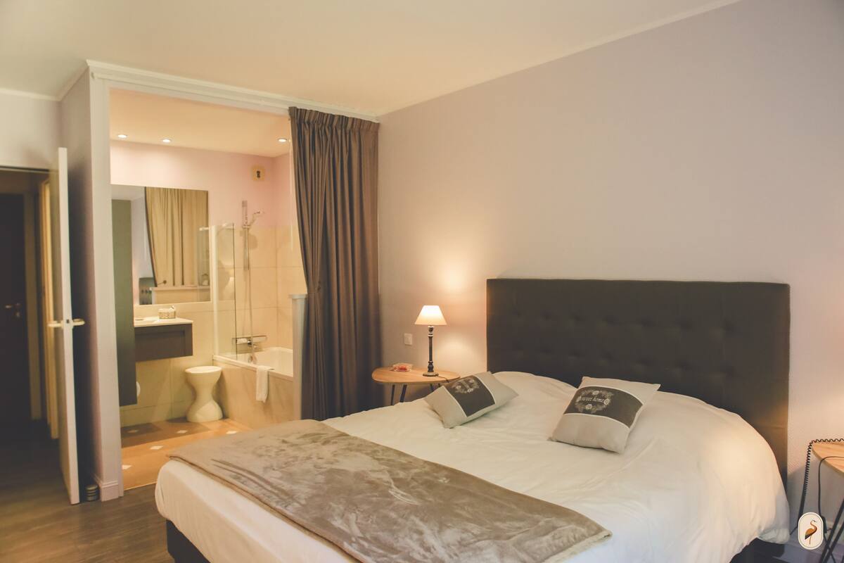 Habitación de hotel con una cama grande, una mesita de noche con lámpara, un baño a la vista a través de una puerta abierta y una zona de ducha.