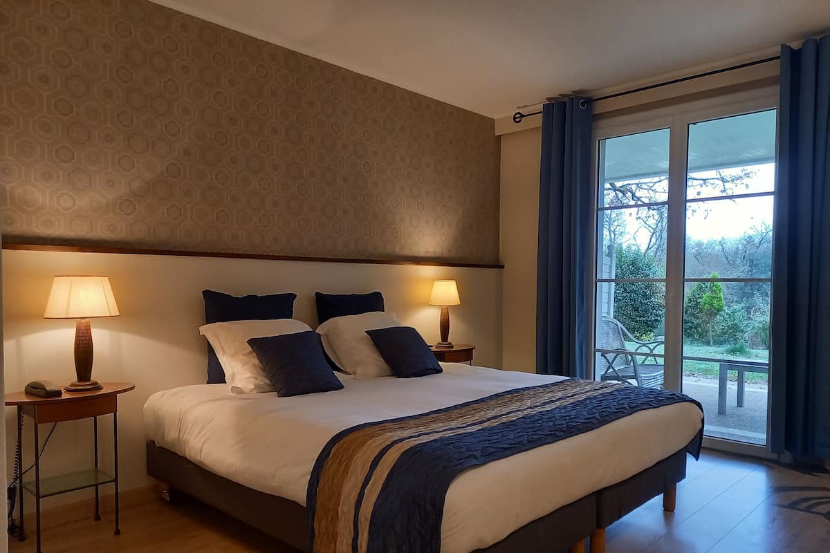 Habitación de hotel con una cama grande, dos mesitas de noche con lámparas, un papel tapiz decorado y vistas amplias por una ventana grande.